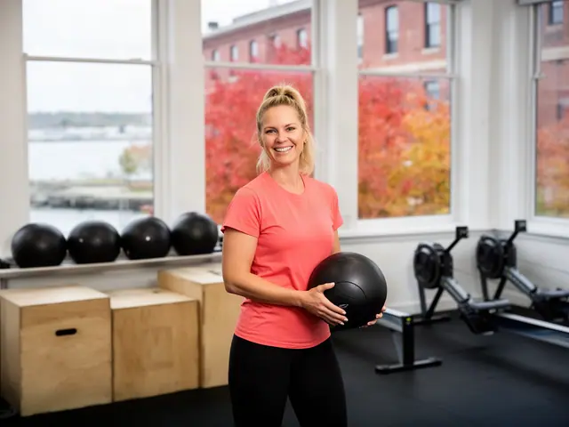 A fitness-studio owner holding a black medicine ball beside plyo boxes and rowing machines.